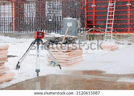 Geodetic device at snow covered construction site in winter season. Optical level at construction site. Equipment for work as surveyor. Optical theodelite on tripod. Red formwork on background Royalty-Free Stock Photo #2120615087
