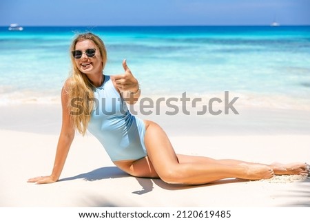 Smiling young woman sitting on the beach and showing thumbs up. She makes recommendations.