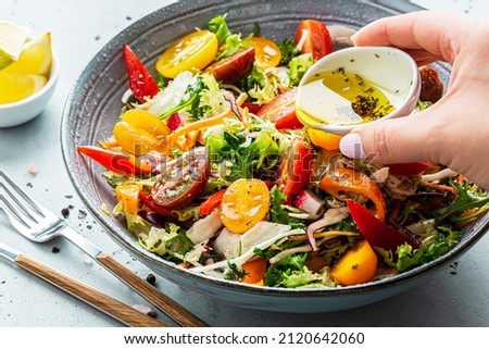 Fresh colorful spring vegetable salad with cherry tomatoes and sweet peppers in the blue bowl. Cook’s hand pouring olive oil with herbs (dressing). Healthy organic vegan lunch or snack close up. Royalty-Free Stock Photo #2120642060