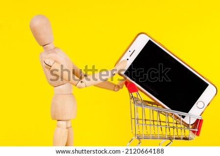 wooden man with a supermarket cart in which a cell phone on a yellow background. High quality photo