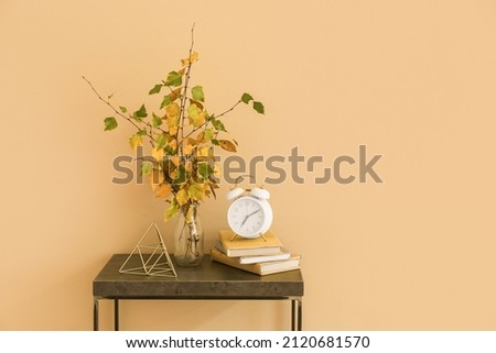 Vase with autumn branches, stack of books and alarm clock on table near color wall