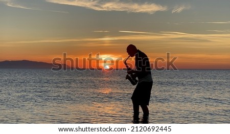 Young male saxophonist stands with his feet in sea water, holds saxophone in his hands, looks at sunset. Beautiful sunset on sea, sky. Musician, playing saxophone, dancing, having fun, Silhouette. Royalty-Free Stock Photo #2120692445