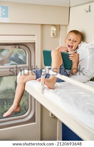 Blond little boy watches cartoons via tablet PC and leaning on soft pillow on top shelf in train compartment in morning