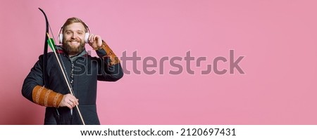 Portrait of young bearded man in image of medieval warrior, archer in chain armor listening to music in headphones isolated over pink background. Comparison of eras, history, renaissance style.