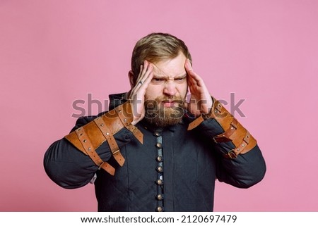 Portrait of young bearded man in image of medieval warrior, archer suffering from pain, touching temples in headache isolated on pink studio background. Comparison of eras, history, renaissance style.