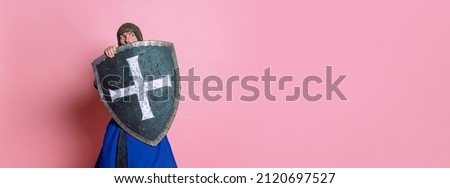 Portrait of scary man, medieval warrior or knight in chain armor hiding himself behind a shield isolated over pink studio background. Comparison of eras, history, renaissance style