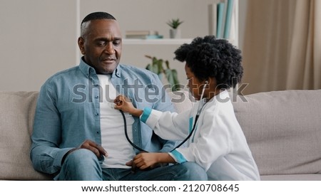 Little african american girl in medical gown plays sitting on couch kid doctor listens to father stethoscope pretends nurse having fun loving daughter forbids dad to smoke indicates get rid bad habit Royalty-Free Stock Photo #2120708645