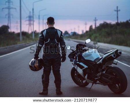 A biker stands next to a motorcycle on an asphalt road and looks into the distance. Summer sunset Royalty-Free Stock Photo #2120719877
