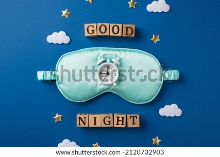 Top view photo of one big silk light blue sleep mask and small white alarm on it with scattered confetti in shape of stars and clouds and wooden cubes with letters on the deep blue background