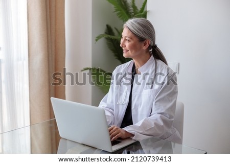 Portrait of happy mature female physician working on laptop in office