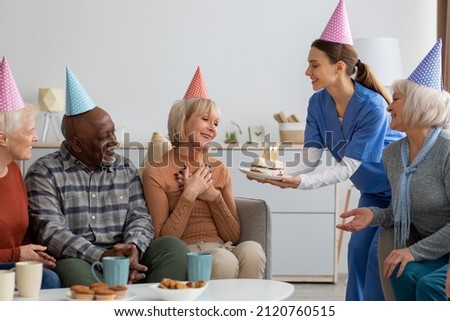 Emotional elderly woman having birthday party at nursing home, sitting on couch among her multiracial friends, looking at birthday cake with lit candles in female nurse hands, wearing colorful hats