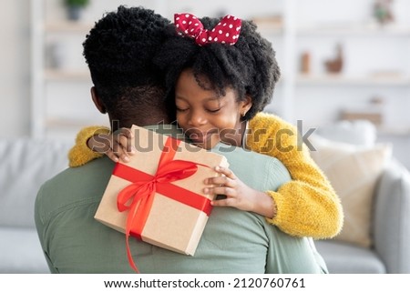 Smiling little black girl holding gift box and embracing dad, loving daughter greeting daddy with Father's day or birthday, happy african american family bonding together at home, closeup shot