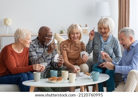 Multiethnic group of happy senior friends men and women in casual sitting on couch, having conversation and playing cards while drinking tea with cookies, chilling together at home Royalty-Free Stock Photo #2120760995