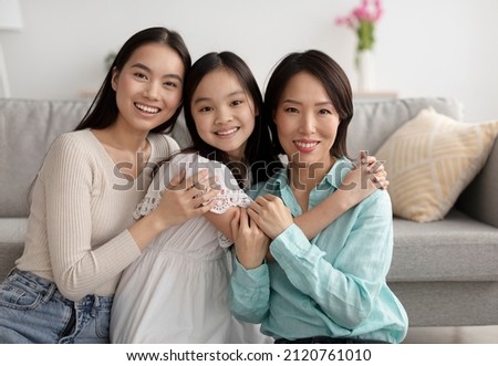 Young Asian woman with her mature mother and adorable little daughter hugging and looking at camera at home. Multi generation family smiling, expressing love and fondness Royalty-Free Stock Photo #2120761010