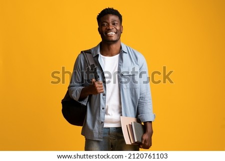 Positive millennial black man student in casual posing with books and notepads on yellow studio background, young african american guy enjoying studying at university or college, copy space Royalty-Free Stock Photo #2120761103