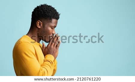 Side view of praying african american young man in yellow, holding palms by his lips and whispering pray, asking for grace over blue studio background, panorama wuth copy space Royalty-Free Stock Photo #2120761220