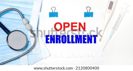 On a light background lie a stethoscope, an electronic thermometer, a syringe, a face mask and a sheet of paper with the text OPEN ENROLLMENT. Medical concept