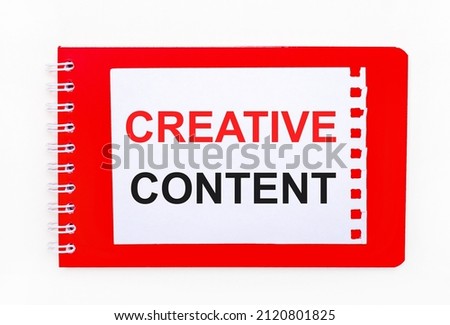 On a white background - a bright red notebook on a spiral. On it is a white sheet of paper with the text CREATIVE CONTENT