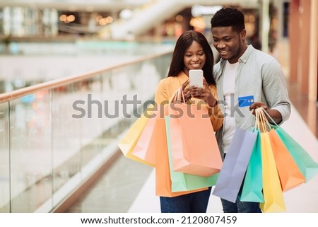 Easy Payment Concept. Portrait of happy African American man and woman using mobile phone together holding credit card and shopping bags, casual couple standing in city mall. Retail And Purchase Royalty-Free Stock Photo #2120807459