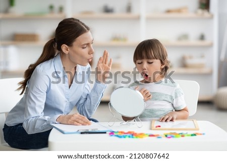 Speech therapy concept. Cute little boy pronouncing sound O looking at mirror, professional woman therapist teaching kid right pronounciation during private lessong at office Royalty-Free Stock Photo #2120807642