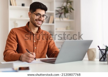 Portrait of young smiling Arab man in glasses using laptop sitting at desk, writing in notebook. Cheerful guy browsing internet, watching webinar studying online, looking at pc screen, free copy space Royalty-Free Stock Photo #2120807768