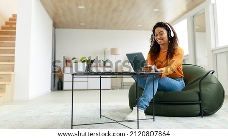 Young smiling black woman holding and using digital tablet sitting at tea coffee table on bean bag chair in living room. Cheerful lady in headset browsing internet listening to music online, panorama