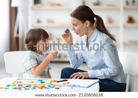 Female speech therapist curing child's problems and impediments. Little boy learning letter O with private English language tutor during lesson at office Royalty-Free Stock Photo #2120808038