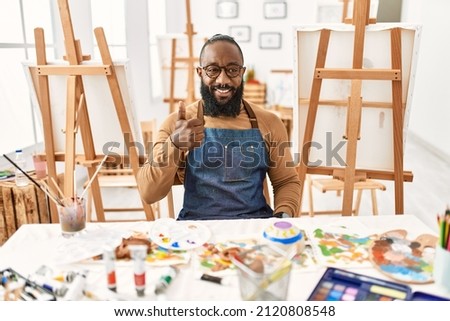 African american artist man at art studio doing happy thumbs up gesture with hand. approving expression looking at the camera showing success. 