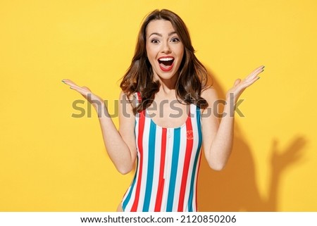 Happy amazed surprised young woman slim body wear striped red blue one-piece swimsuit spreading hands isolated on vivid yellow color wall background studio. Summer hotel pool sea rest sun tan concept