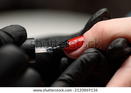 Manicurist makes a hardware manicure to a client of a beauty salon.
The master applies a decorative bright red varnish on the nail plate with a brush. 
The concept of beauty and nail care.