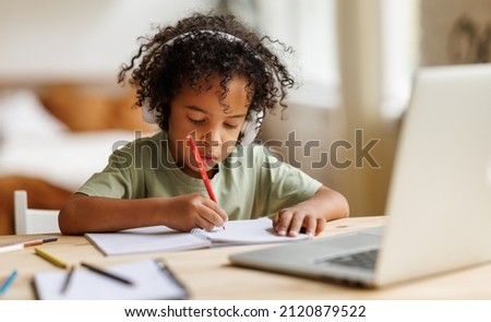 Distance education. Smiling african american child schoolboy in headphones studying online on laptop at home, sitting at table and communicating with teacher through video call on computer