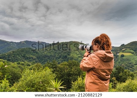 Young unrecognizable Caucasian woman in an orange coat taking photos in the middle of the mountains in the middle of nature on a cloudy day