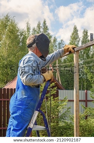 Man welder in a welding mask, construction uniform and protective gloves cooks metal on a street construction site. Construction of a pavilion, pergola near a country house on a summer day