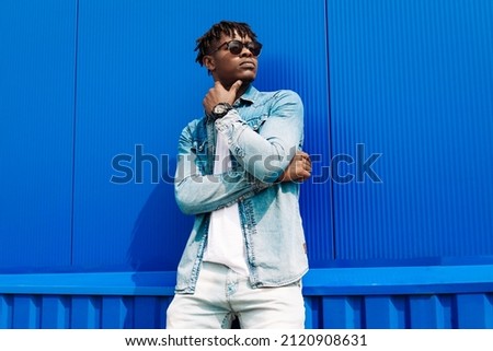 Fashion portrait of a handsome smiling stylish dark-skinned man, dressed in a jacket and jeans. Fashionable man posing against blue wall in sunglasses Royalty-Free Stock Photo #2120908631