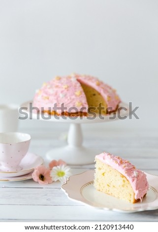 Pink frosted cake on a plate stand with a cake slice and tea cup and flower decorations on a white wood table