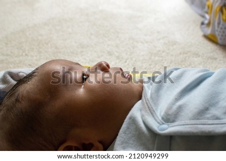 A Japanese Asian infant baby with blue clothes lying down on the white carpet looks at his mother and gets relaxed