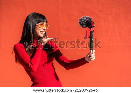 Pretty brunette girl giving a kiss and recording a video blog with the mobile phone and microphone, wearing a dress for social networks. Caucasian pretty a red background, copy space and paste
