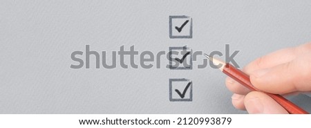 hand with pencil writing check mark on Checklist. Hand with pencil and check boxes on gray paper background. Male hand choosing three of three options. Royalty-Free Stock Photo #2120993879