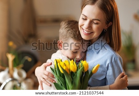 Little son congratulating mom with Mothers day at home, embracing her and giving her fresh flower bouquet, happy   woman mother embracing son. Family holidays and celebration concept