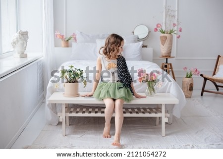 A beautiful little girl with curly hair in a green skirt sits in a spacious bright room among the flowers. Delicate photo. The child poses in the bedroom by the big white bed. Vase with pink tulips.