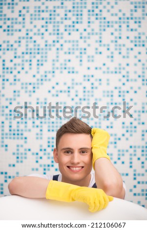 Half-length portrait of young dark-haired smiling janitor wearing white T-shirt blue overalls and yellow rubber gloves leaning on the wash bowl in the bathroom dreaming about new job
