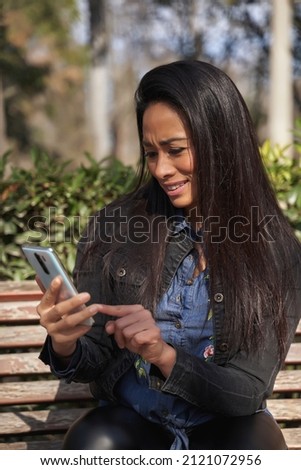 Young woman sitting on a bench in a green park on a spring day and reading a text message on the cell phone. High quality photo