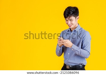 Portrait studio shot of millennial Asian professional successful male businessman employee in formal outfit standing smiling holding  smartphone  playing virtual video game on yellow background.