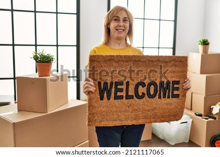 Middle age blonde woman smiling happy holding doormat at new home.