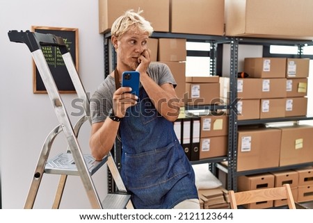 Young blond man using smartphone working at storehouse looking stressed and nervous with hands on mouth biting nails. anxiety problem. 