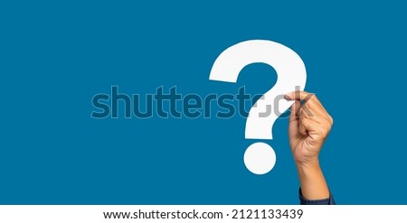 Hand holding a white question mark paper against a blue background in the studio. Question mark symbol concept. Close-up photo. Space for text.
