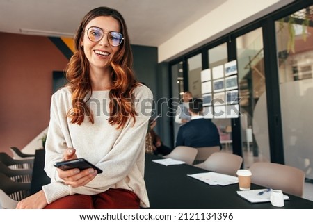Happy young businesswoman smiling at the camera while holding a smartphone. Cheerful young businesswoman sitting on a boardroom table with her colleagues in the background.