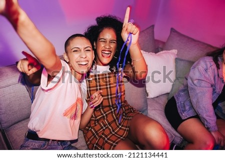 Two female friends taking a selfie at a house party. Two happy young women taking a picture together while sitting on a couch in neon light. Best friends having a good time together on the weekend.