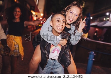 Playful young woman piggybacking her best friend outdoors at night. Happy young women having fun while going out with their friends in the city. Vibrant friends spending their weekend together. Royalty-Free Stock Photo #2121134453