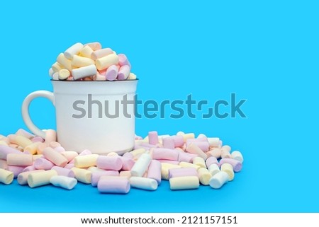 A mug with cocoa or hot chocolate and a lot of colorful marshmallows scattered on a blue background. Sweet airy dessert or addition to hot drinks. Marshmallows with cocoa and free space for text